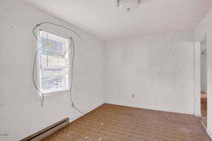 An empty room with white walls shows visible stains and marks. The floor has a brown checkered pattern and debris scattered. A single window with blinds and loose hanging cords is on the left, and a ceiling light is above.
