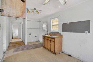 A small, worn kitchen with a double sink, wooden cabinets, and peeling linoleum flooring. There are missing appliances, exposed wiring, damaged walls, and an open hallway leading to other rooms.