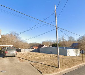 A utility pole with overhead wires stands near a driveway where an SUV is parked. The area has patchy grass, a metal building, wooden fences, and several houses with trees in the background under a clear blue sky.