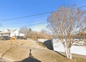 A grassy, sloped lot with a sidewalk runs alongside a white fence and bare tree at 1 Mcclanahan Street, Oxford. A house and parked car are visible on the left, with overhead wires and a yellow street sign under a clear blue sky.