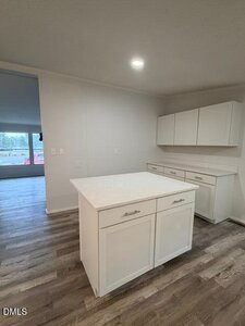 A modern kitchen with white cabinets and a central island, featuring a neutral color palette and wood-look vinyl flooring. A bright living area is visible through an open doorway in the background.