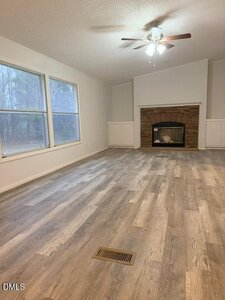 A spacious living room with light wood flooring, large windows on the left, a ceiling fan with light, and a brick fireplace with built-in white cabinets on either side. The room is empty and well-lit.