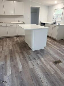 A modern kitchen with white cabinets, a white center island, stainless steel fixtures, light gray walls, and wood-look laminate flooring. A window is above the sink and an open pantry is visible in the background.