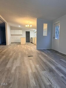 A bright, empty room with light wood-style flooring, white walls, and a view of a kitchen area with white cabinets and an island. A door with a window is on the right side. A ceiling light fixture hangs above.