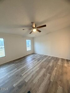 An empty room with light-colored walls, wood-look vinyl flooring, two tall windows, and a ceiling fan with a light fixture in the center of the ceiling.
