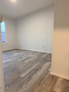 An empty room with light gray walls, a window on the left, and wood-style vinyl flooring. The scene is well-lit with natural and artificial light.