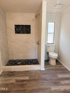 A bathroom with a walk-in shower featuring large beige tiles, a black hexagonal tile floor and niche, a white toilet next to a window, and wood-look vinyl flooring.