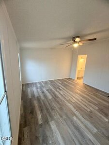 A mostly empty room with light-colored walls, wood-look laminate flooring, a ceiling fan with a light, and a small doorway leading to a bathroom vanity area. Two windows are visible on the left wall.