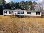A single-story manufactured home at 1185 Diamond Hills Road, Stem, with white siding, black shutters, and a brick foundation. Wooden steps lead to the front door. The house sits on a grassy lot with trees under a partly cloudy sky.