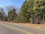 A paved road with double yellow lines, known as 00 Webb Chapel Road, runs alongside a forested area near Oxford, where pine and leafless trees stand under a clear blue sky and dry leaves cover the ground near the edge of the woods.