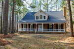 A brick house with a large front porch, white railings, and dark shutters sits in a pine forest. The roof is dark gray, and the yard is covered with dry pine needles. The area is surrounded by tall trees.
