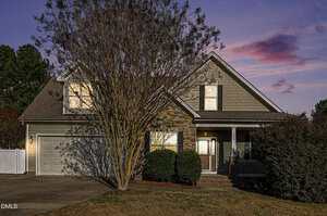 A two-story suburban house with stone and shingle siding, a covered front porch, attached two-car garage, and a large tree in the front yard. The photo is taken at dusk with a pink and purple sky.