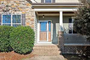 Front entryway of a house with a brown wooden door, glass storm door, brick steps, covered porch, column, stone and siding exterior, bushes on the left, and a railing with vertical bars on the right.