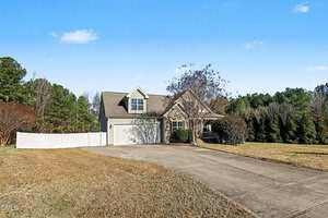 A single-story house with a gable roof, dormer window, and attached two-car garage sits at the end of a wide concrete driveway. The yard is spacious with trees and a white fence on one side. The sky is clear and blue.
