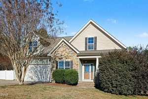 A single-story house with stone and siding exterior, gabled roof, double front windows, a covered entryway, and bushes and trees in the yard under a clear blue sky.