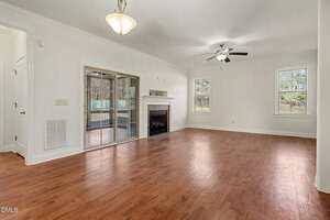 A spacious, empty living room with hardwood floors, a ceiling fan, two windows, a fireplace, and sliding glass doors leading to an outdoor area. Light fixtures hang from the ceiling.