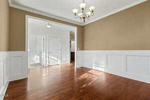 A dining room with wooden floors, beige upper walls, white wainscoting, a brass and glass chandelier, and an open entryway leading to a hallway with white doors and natural light.