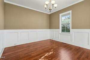 An empty room with beige walls, white wainscoting, a window with outside view, hardwood floors, and a ceiling light fixture with multiple bulbs.