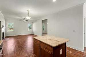 A spacious, empty open-concept living area with hardwood floors, white walls, and large windows. A kitchen island with a granite countertop and brown cabinets is in the foreground. Ceiling fan and light fixtures are visible.