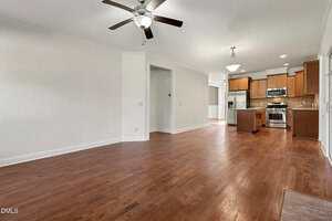 Spacious open-plan room with wood floors, white walls, ceiling fan, and overhead lights. The kitchen in the background features wooden cabinets, stainless steel appliances, and a tile backsplash.