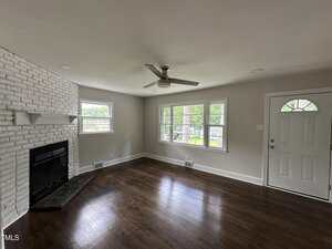 A living room with dark wood floors, a white brick fireplace, light gray walls, three windows, a ceiling fan, and a white door with a window. The room is empty and receives natural light.