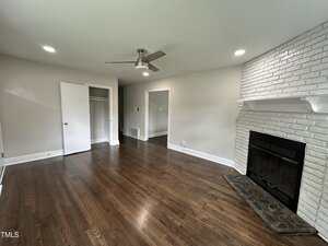 A room with hardwood floors, white walls, recessed ceiling lights, a ceiling fan, an open closet, and a white painted brick fireplace with a mantle. The room is empty and well-lit by natural and artificial light.