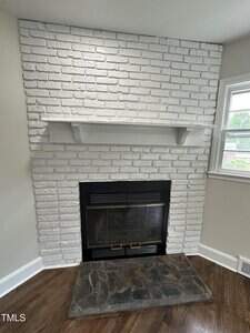 A white-painted brick fireplace with a black metal insert and glass doors, topped by a simple white mantel shelf. The hearth is a slab of dark stone, and a window is visible to the right.