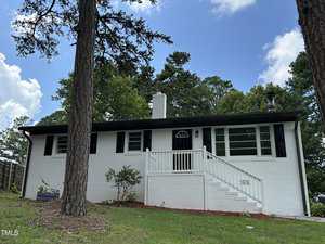 A white brick, single-story house with black shutters and trim, a central chimney, and a front porch with white railings. Stairs lead up to the porch, and tall pine trees are in the yard. The sky is partly cloudy.