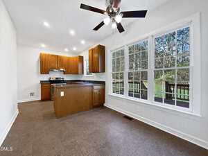 A kitchen with wooden cabinets, stainless steel appliances, an island, and a ceiling fan next to a bright dining area with large windows overlooking a deck and wooded backyard.