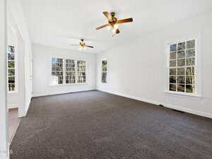 A spacious, empty room with gray carpet, white walls, two ceiling fans with lights, and several large windows allowing natural light to enter. Trees are visible outside the windows.