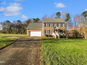 A two-story brick house with blue shutters and a white front porch stands at the end of a wide driveway, surrounded by a large, well-kept lawn and trees under a clear blue sky.