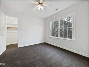 A bright, empty room with white walls, dark carpet, a ceiling fan with lights, a window overlooking trees, and an open door leading to a walk-in closet with a shelf and hanging rod.