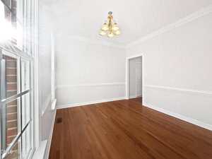 A bright, empty room with white walls, crown molding, hardwood floors, a window on the left, a gold chandelier on the ceiling, and a doorway leading to another room.