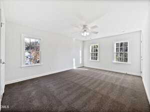 A spacious, empty room with white walls, three large windows, a ceiling fan with lights, and brown carpeted flooring. Natural light enters through the windows, and the room appears clean and well-lit.