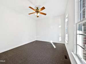 A bright, empty room with white walls, a brown carpet, a ceiling fan with lights, and large windows allowing natural light to enter.