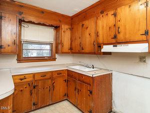 A small, dated kitchen with wood-paneled cabinets, white countertops, a single-basin sink, a window with blinds, and an older range hood. The floors are vinyl tile and the walls show some signs of wear.