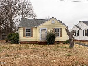 A small, single-story yellow house with white trim and brown shutters, a central front door, brick steps, and a bare tree stump in the front yard. The grass is dry and trees without leaves are in the background.