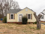 A small, single-story yellow house with white trim and brown shutters sits at 319 E Church Street in Creedmoor. A bush grows in front of the door, with a bare tree trunk in the yard and leafless trees beneath a cloudy sky in the background.