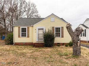 A small, single-story yellow house with white trim and brown shutters sits at 319 E Church Street in Creedmoor. A bush grows in front of the door, with a bare tree trunk in the yard and leafless trees beneath a cloudy sky in the background.