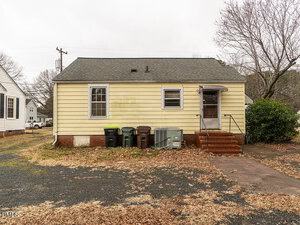 A small yellow house with a gray shingled roof and red brick steps leading to a back door. Four trash bins and two HVAC units are lined up against the exterior wall. The yard has scattered leaves and a partially paved surface.