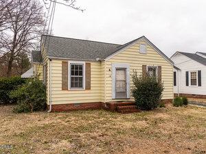 A small, single-story yellow house with brown shutters, a white front door, and a brick foundation. There is a bush near the entrance and patchy grass in the front yard. The sky is overcast.