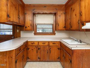 A kitchen with wood cabinets, white countertops, a single basin sink, a window with blinds above the sink, and a beige tiled floor. The cabinets have visible knots and black handles.