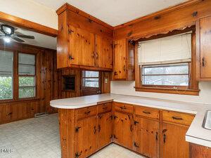 A kitchen with wood-paneled walls and cabinets, white countertops, a window with blinds above the sink, and a door leading outside. The floor has light-colored linoleum tiles.