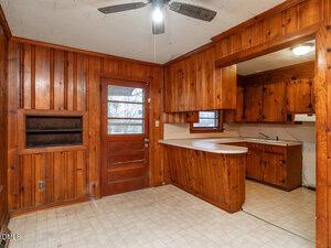 A kitchen with wood-paneled walls and cabinetry, a ceiling fan with a light, a back door with windows, a small counter dividing two areas, vinyl flooring, and a corner sink under a window.