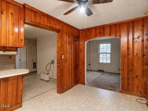 A room with wood-paneled walls, a ceiling fan, and an open doorway leading to a carpeted area with a window. Another opening leads to a kitchen with light-colored flooring and cabinets.