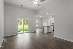 A spacious, empty room with light wood flooring, white walls, a ceiling fan, sliding glass doors leading to a backyard, and an open view into a modern kitchen with white cabinets and recessed lighting.