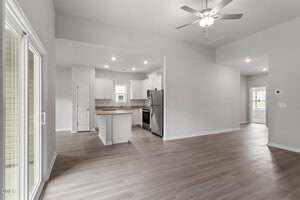 A modern, empty open-plan living area with light wood flooring, a ceiling fan, and neutral walls. The kitchen has white cabinets, stainless steel appliances, and a small island. Sliding glass door and windows provide natural light.