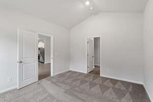 A clean, empty room with light gray carpet, white walls, and a high ceiling. Two open doors show adjoining rooms; one leads to a hallway with wood flooring and kitchen appliances visible.