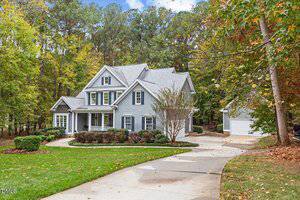 A two-story gray house with white trim sits at the end of a curved driveway, surrounded by trees with autumn foliage. There is a detached white garage in the background and a landscaped front yard.