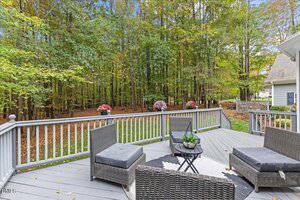 A gray wooden deck with wicker patio furniture overlooks a grassy backyard bordered by dense trees. Pink flowers decorate the railing, and neighboring houses are partially visible.